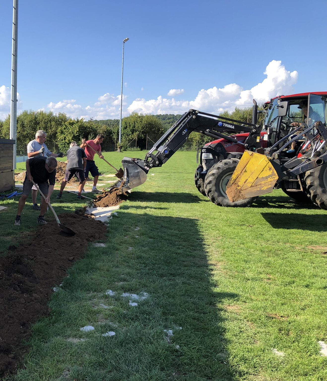 Vereinsmitglieder legen mit Traktor und Schaufeln den Graben für die Bewässerung am Spielfeldrand an